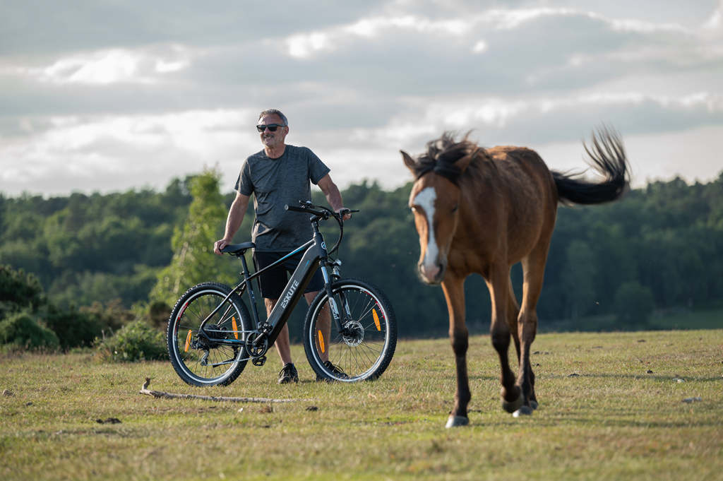 En man står bredvid en häst och håller fast sin elektriska mountainbike med kedjedriven växel