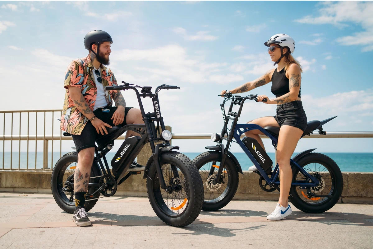 A man and a woman with black and blue V202 retro electric bikes chatting on a sunny beach broadwalk.