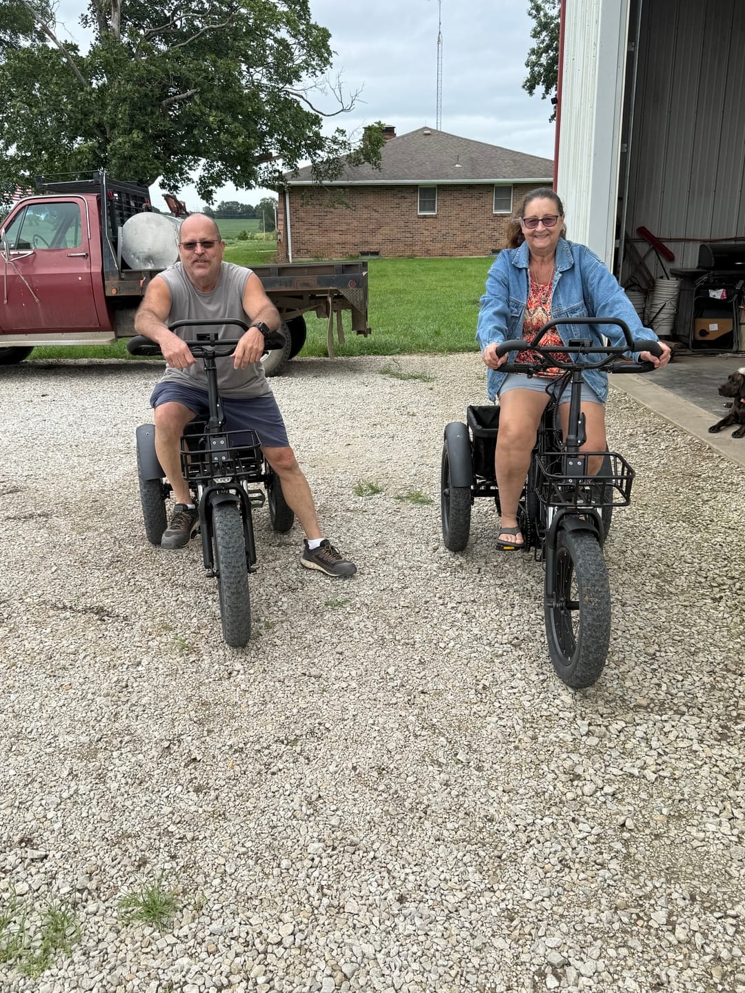 Customer's two people sitting on black Eskute T302 Pro Cargo E-Bikes on a gravel driveway near a truck and building.