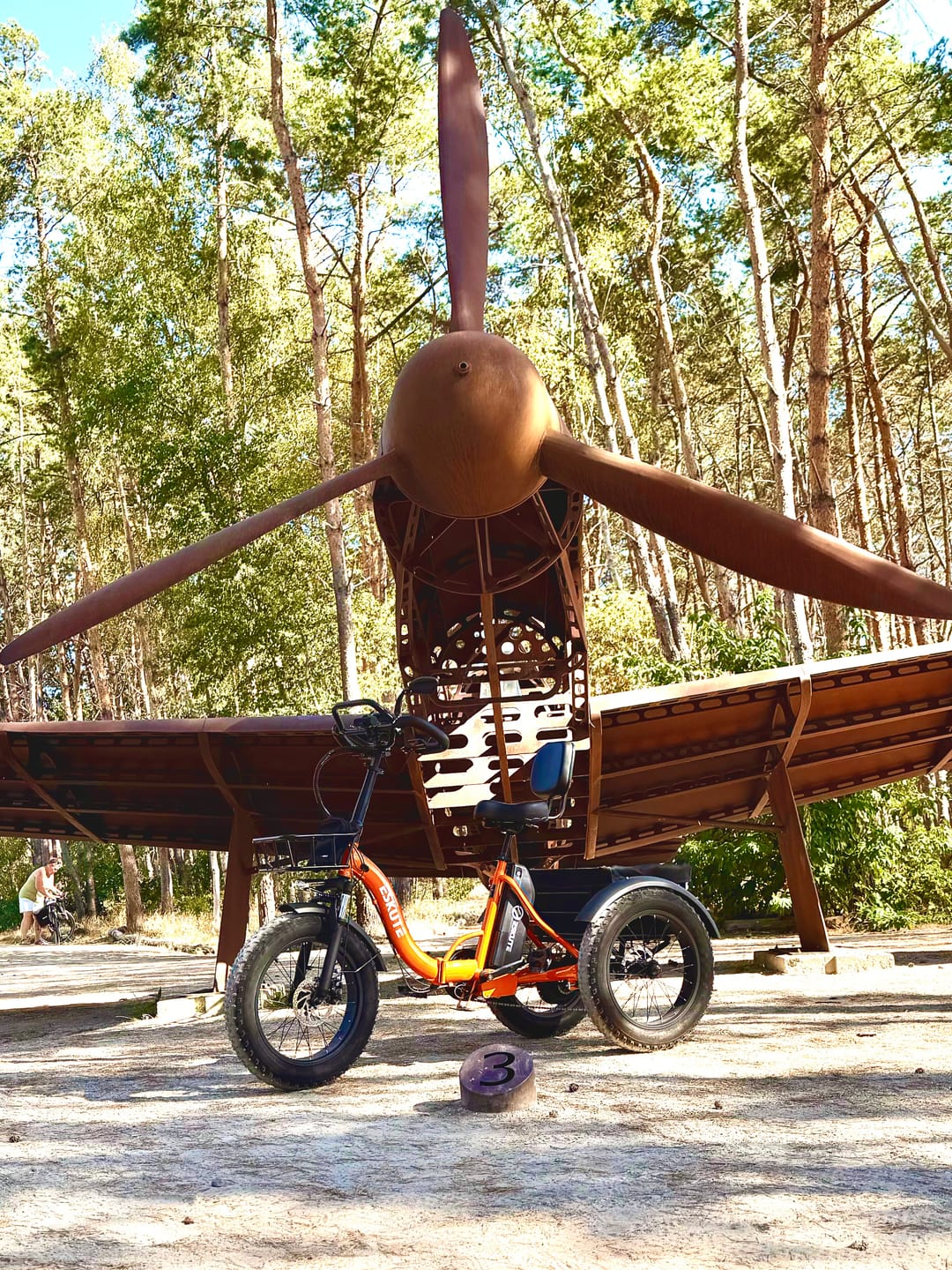 Customer's orange Eskute T302 Pro Cargo E-Bike parked on a sandy path in front of a large metal airplane sculpture.