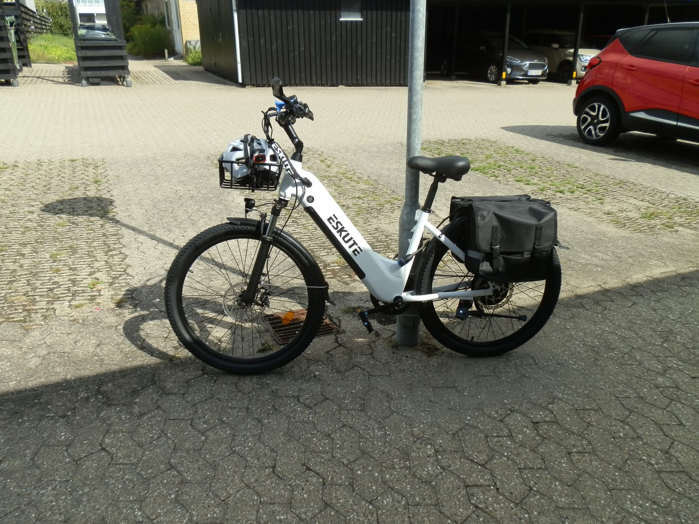 Customer's white Eskute C202 City E-Bike parked next to a pole, featuring a front basket and rear storage bag.