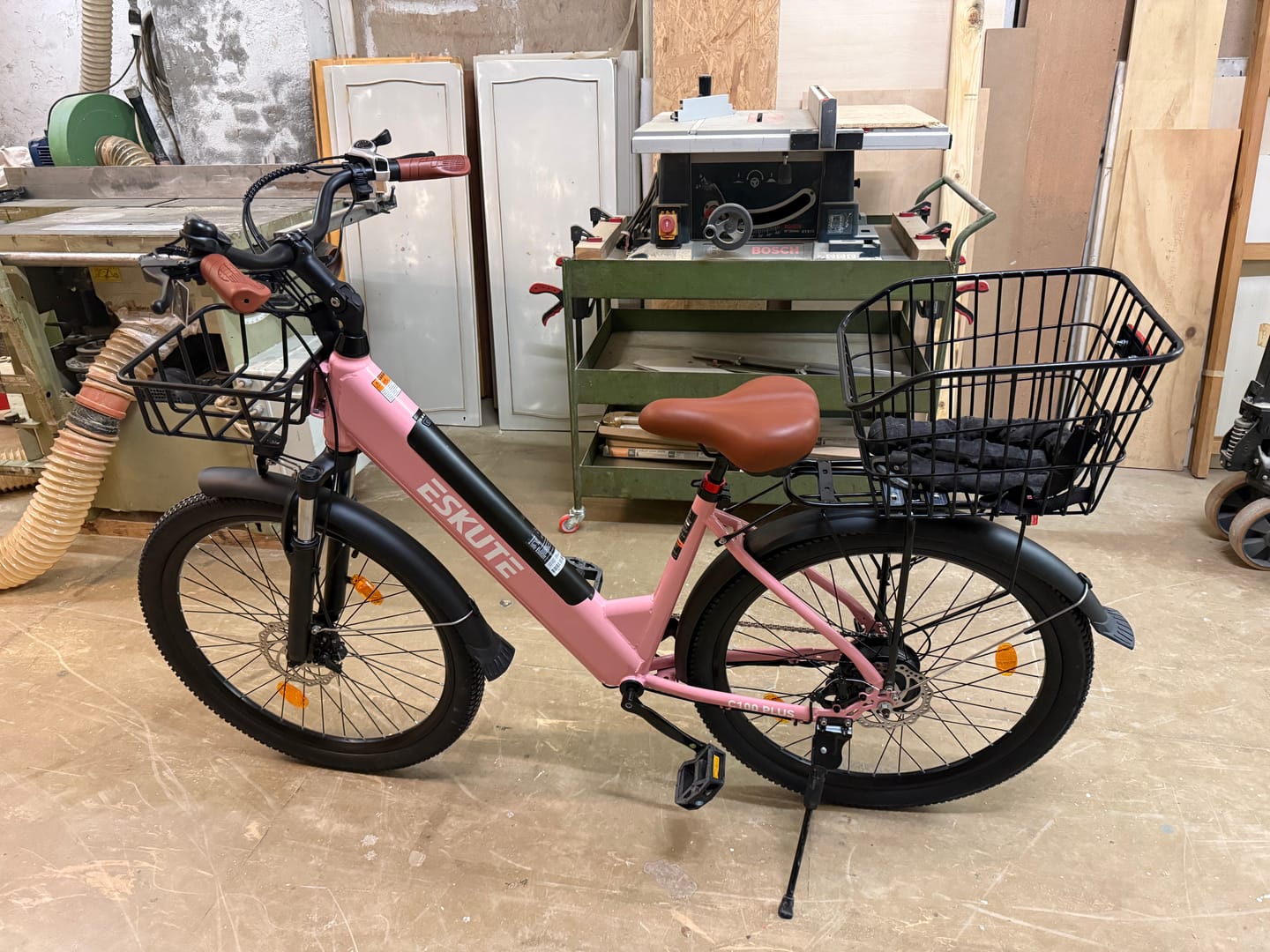 Customer's pink Eskute C100 Plus City E-Bike with front and rear wire baskets, set up inside a workshop.